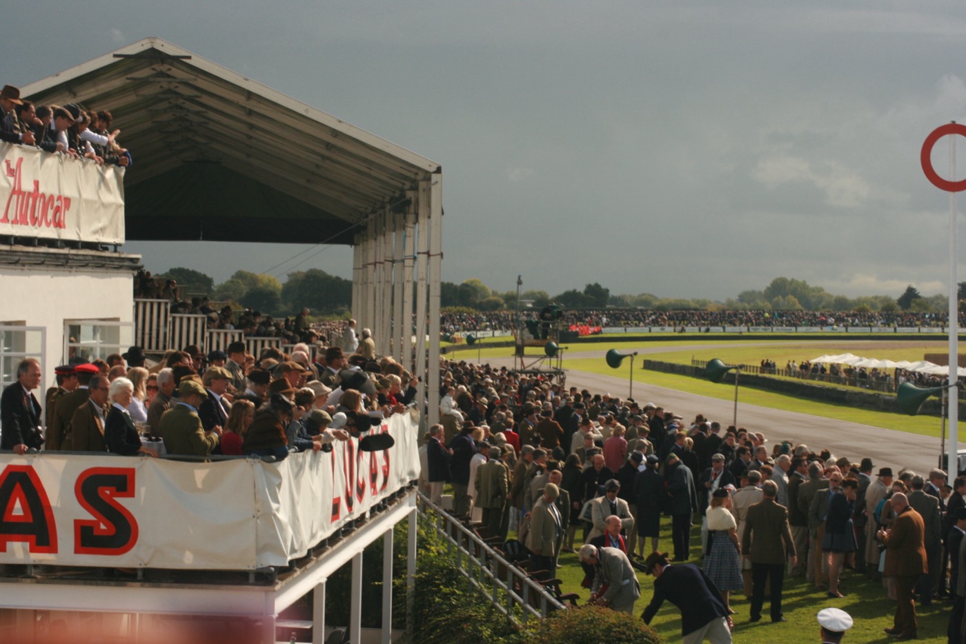 Crowd in tweed jackets, trilbies, flat caps and 1950s dresses beside the track and on a grandstand balcony at the Goodwood Revival