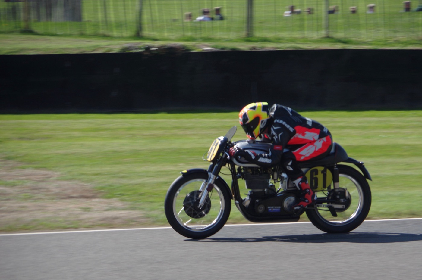Rider in black and red leathers crouched over a black Manx Norton racing motorcycle, number 61, on the Goodwood circuit