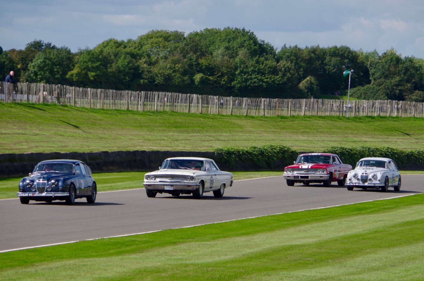 A dark blue Jaguar Mk2, two Ford Galaxie 500s and a white Jaguar Mk2 racing side by side on the Goodwood circuit, with a grass bank and wooden fence behind