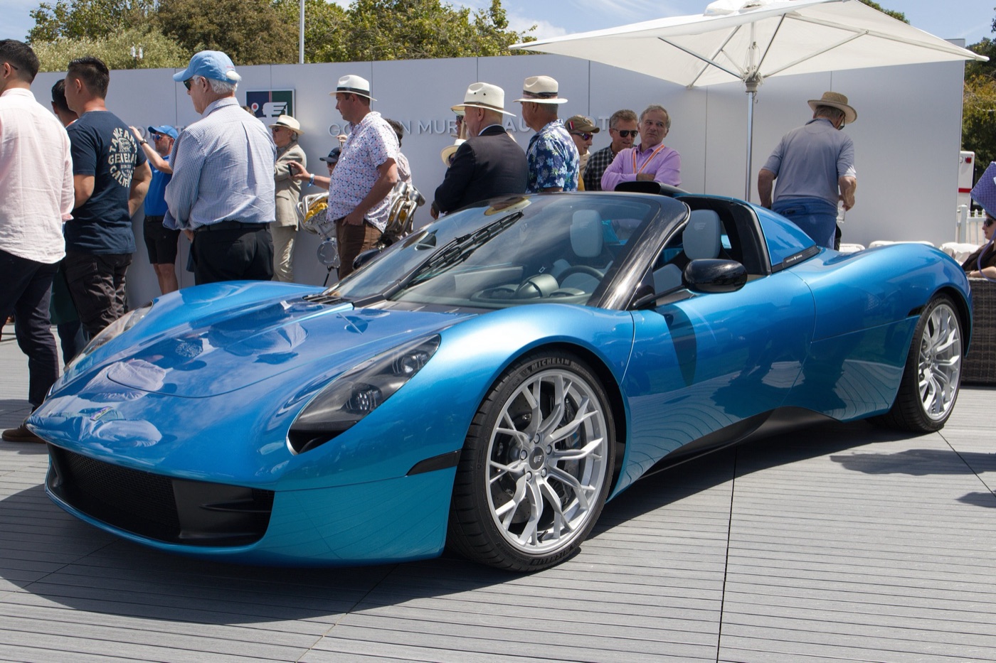 Bright blue Gordon Murray Automotive T.33 Spider with its roof panels removed, on a display stand with visitors behind