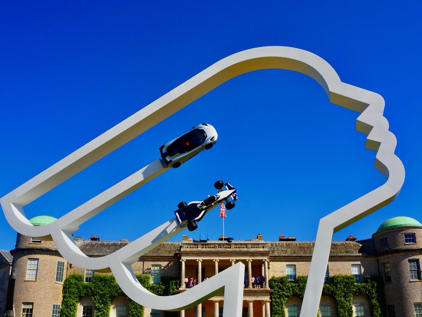 White outline sculpture in front of Goodwood House carrying a Brabham BT52 and a silver Gordon Murray supercar high in the air