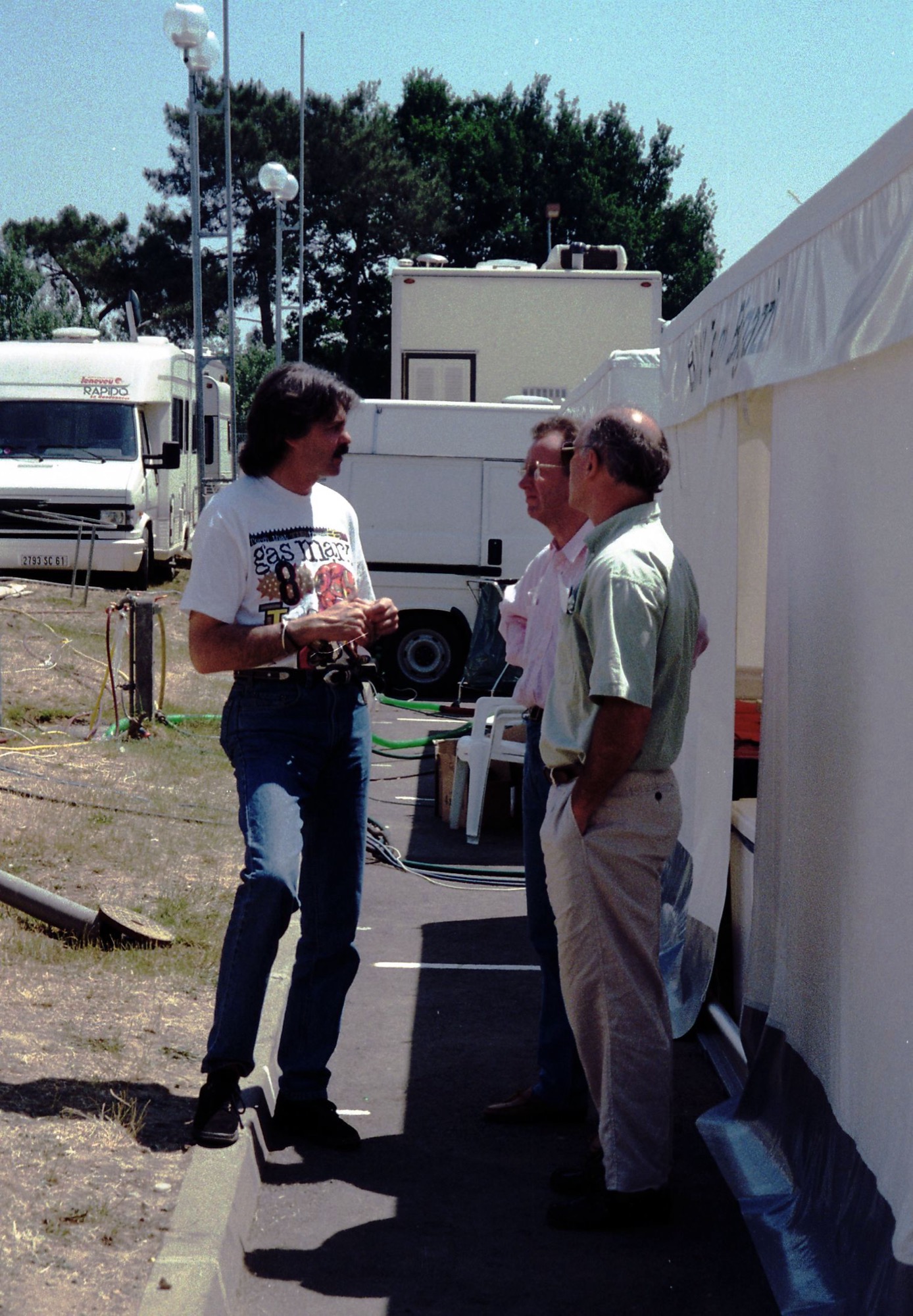 Gordon Murray, long-haired in a T-shirt and jeans, talking to two men in the paddock behind the pits at Le Mans