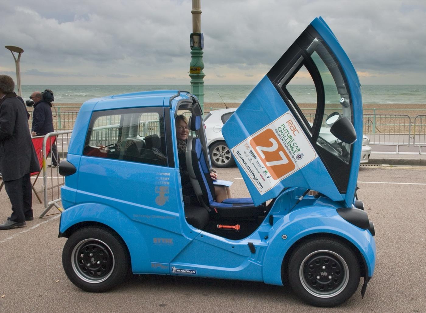 Light blue Gordon Murray Design T.27 electric city car with its front-hinged door raised, wearing RAC Future Car Challenge number 27 on Brighton seafront
