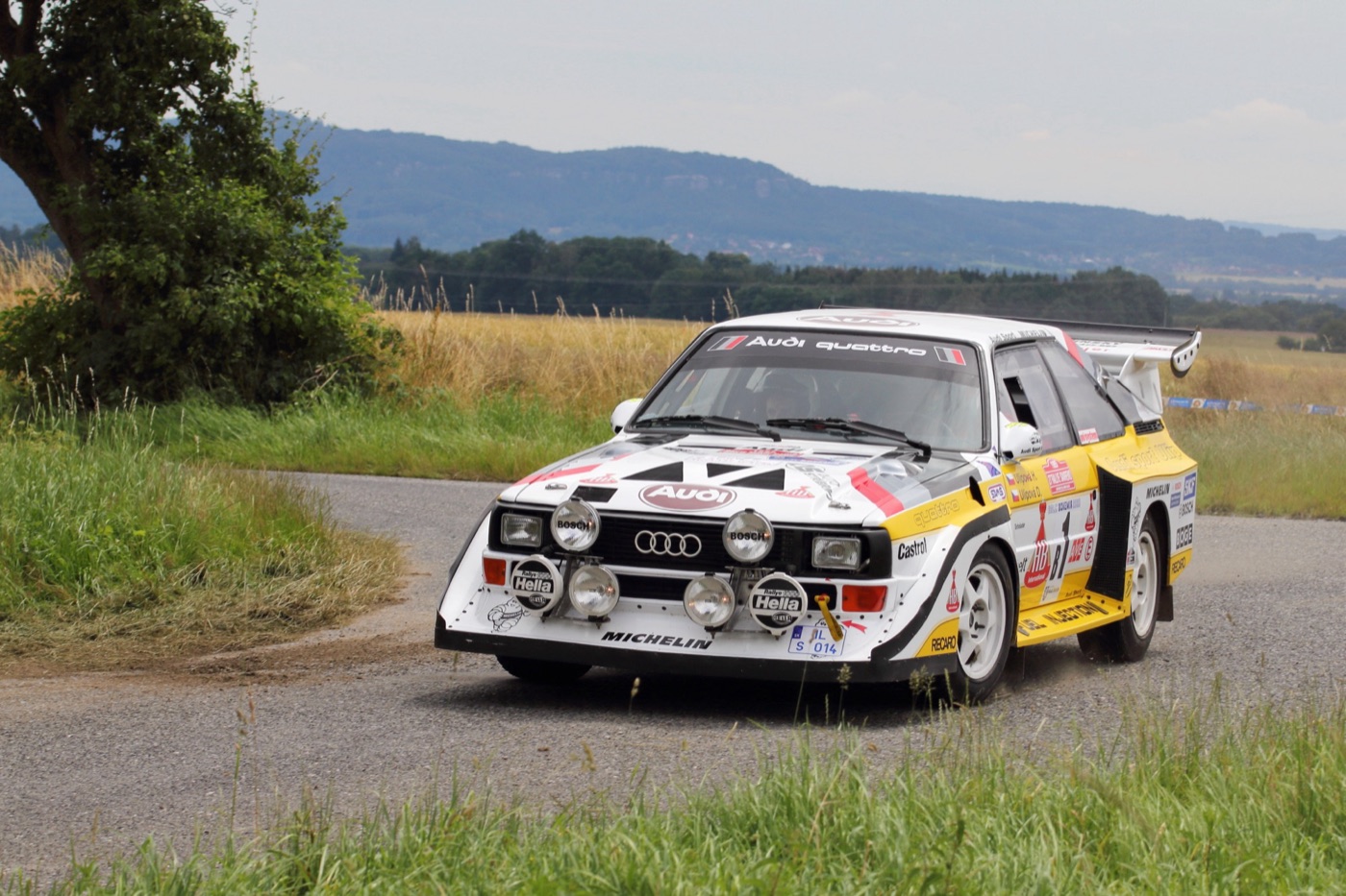 White, red and yellow Audi Sport quattro S1 E2 rally car with a large rear wing, cornering on a country tarmac stage with hills behind