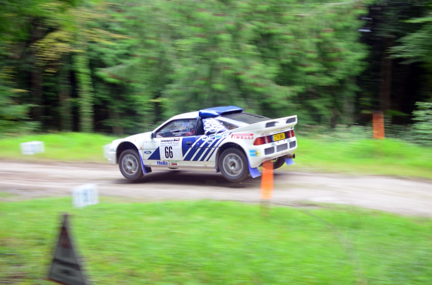 White and blue Ford RS200 rally car, number 66, airborne over a crest on a forest gravel stage