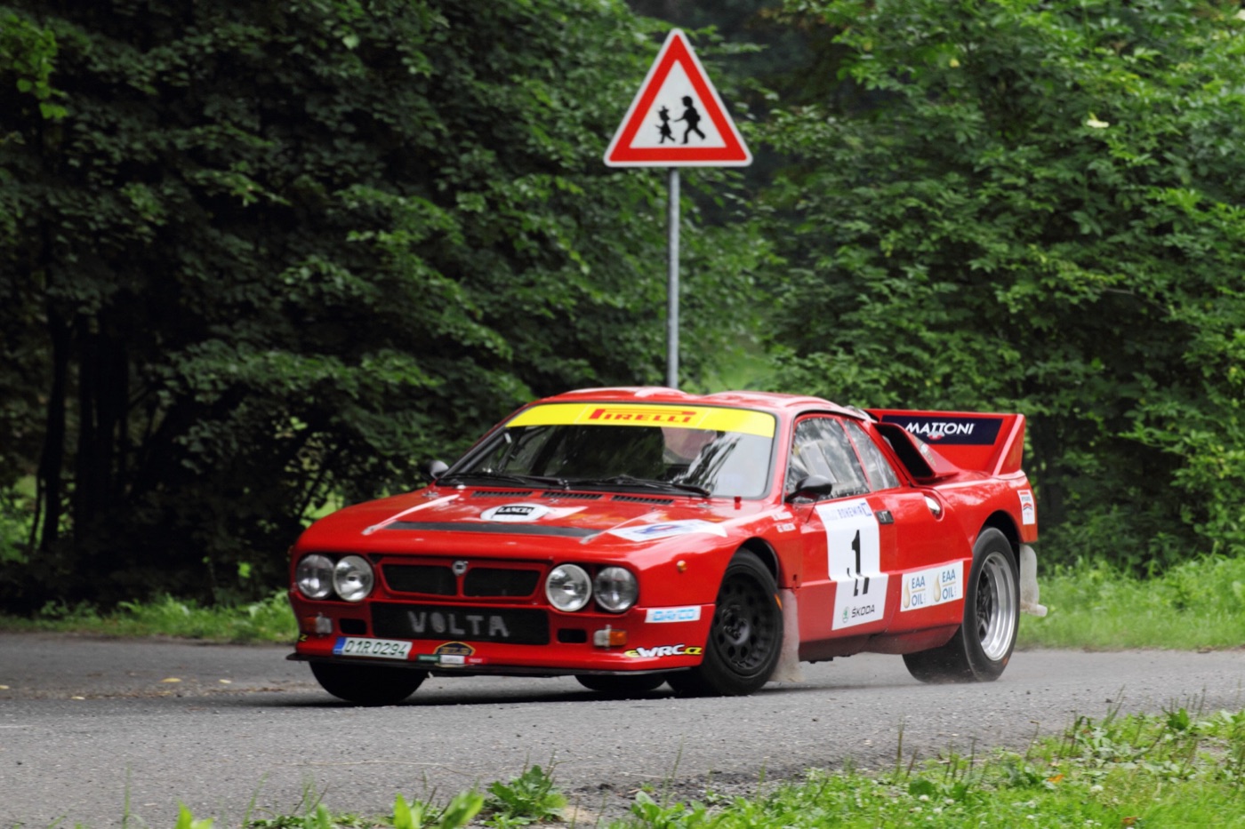 Red Lancia 037 rally car with a yellow Pirelli windscreen band, number 1, cornering on a tree-lined tarmac road