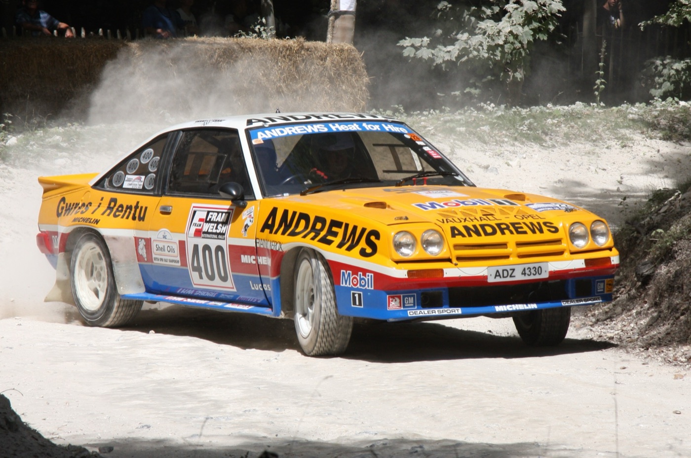 Yellow, red and blue Opel Manta 400 rally car, number 400, in Andrews Heat for Hire livery, sliding on a dusty gravel stage