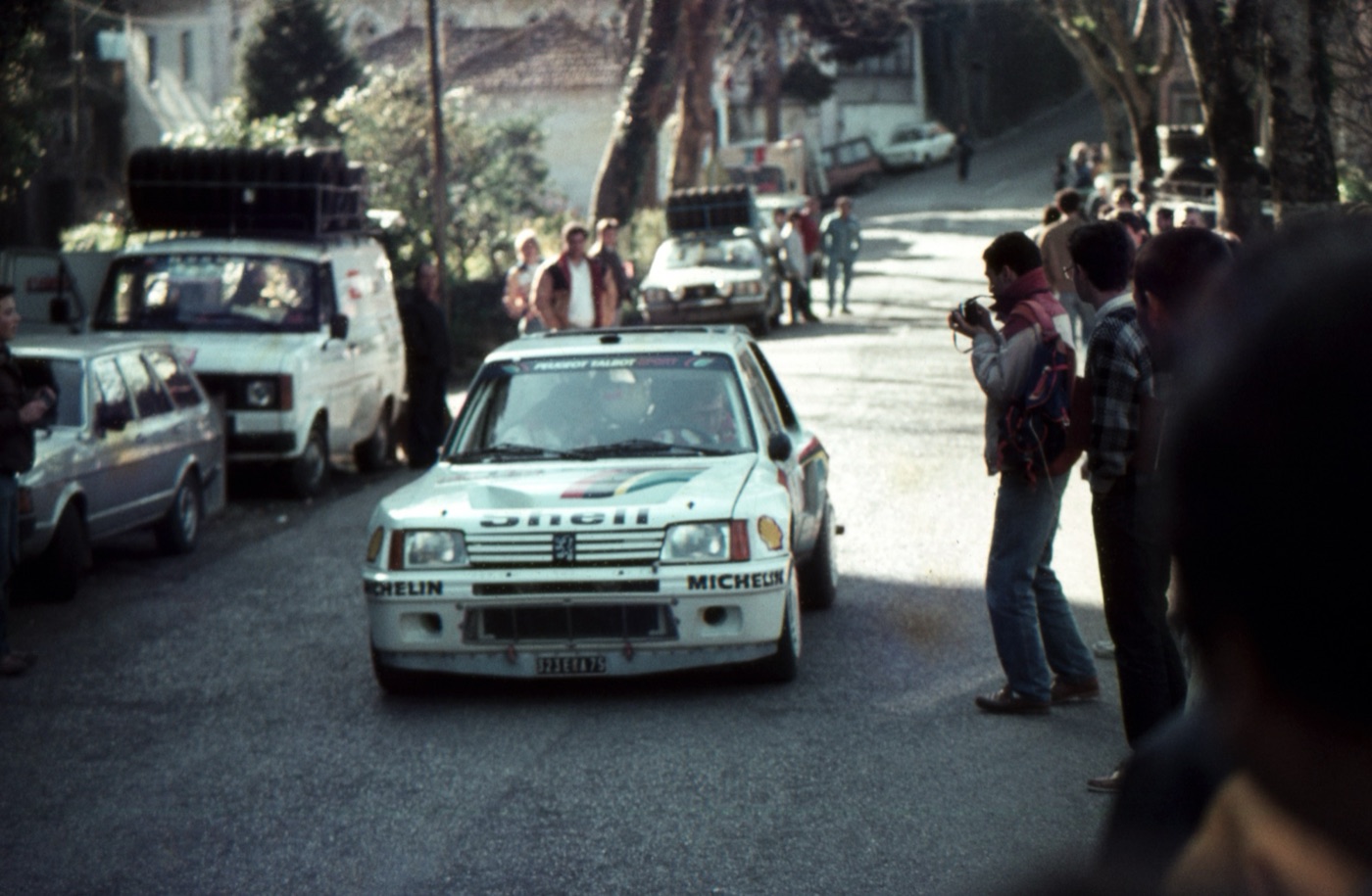 White Peugeot 205 Turbo 16 rally car driving slowly along a narrow town street lined with spectators and service vans