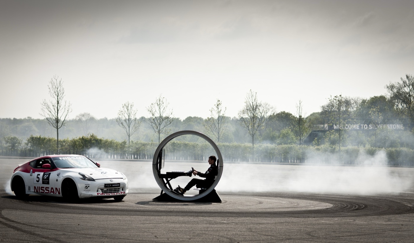 A Nissan 370Z in GT Academy livery drifts in a circle round a driver in a ring-shaped PlayStation gaming pod, with a Welcome to Silverstone sign behind