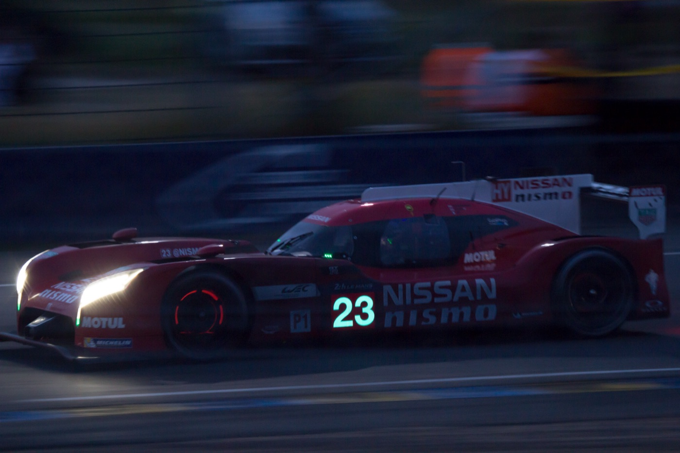 Red Nissan GT-R LM Nismo LMP1 car, number 23, at dusk with its headlights on and glowing brake discs