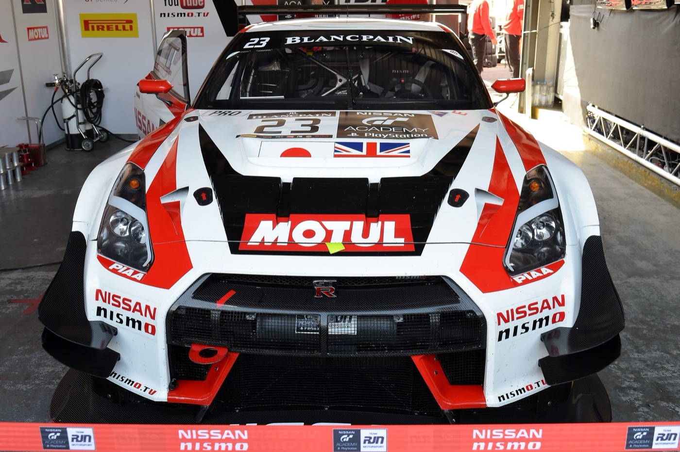 Front of a white and red Nissan GT-R Nismo GT3, number 23, with a GT Academy PlayStation panel on its bonnet, in a pit garage