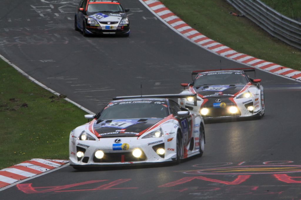 Two white, black and red Gazoo Racing Lexus LFAs, numbers 51 and 50, cornering with headlights on at the Nürburgring