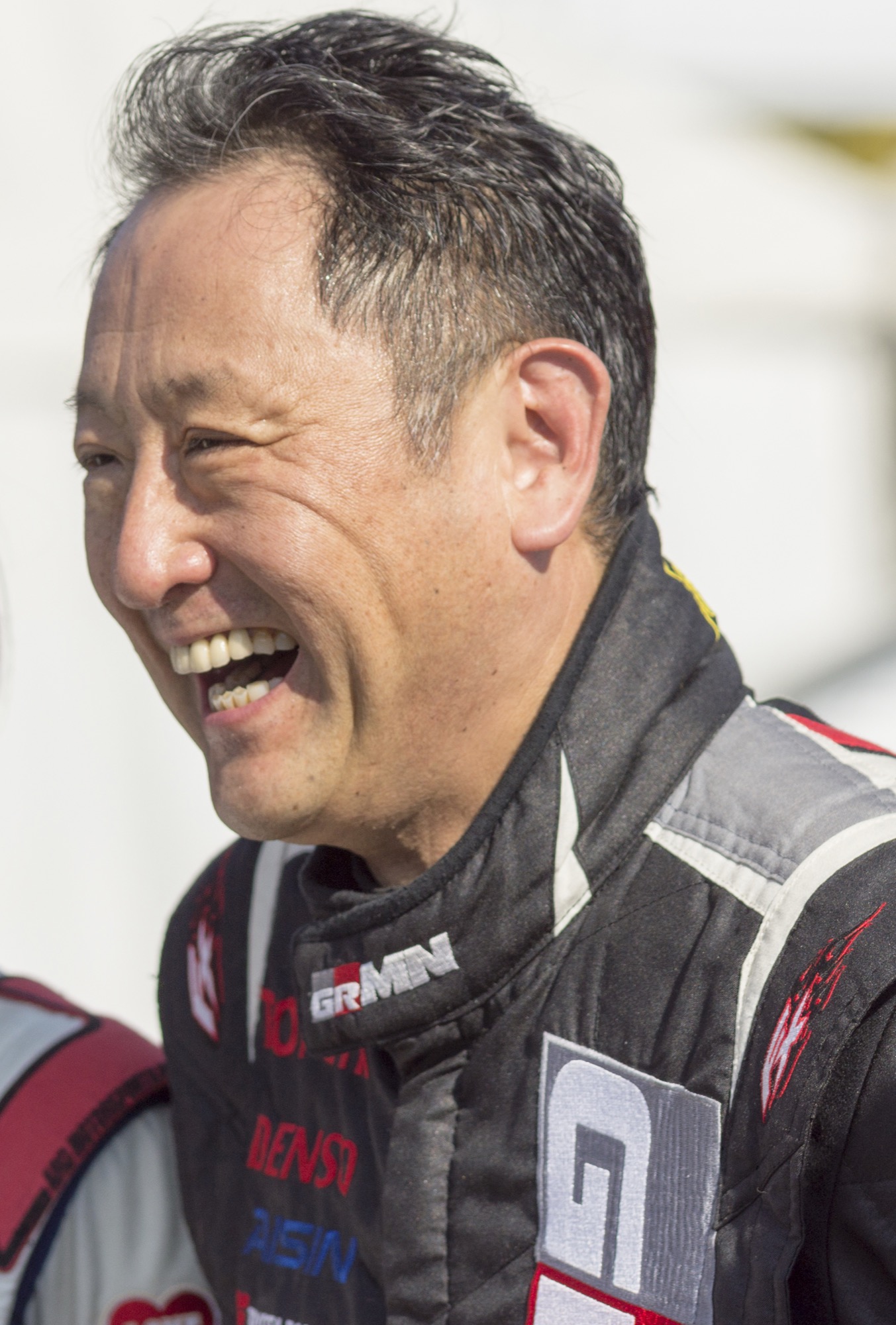 Akio Toyoda laughing in a black Gazoo Racing race suit with a GRMN badge on the collar
