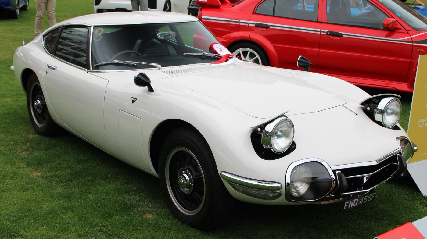 White Toyota 2000GT coupe with pop-up and fixed headlamps parked on grass at a concours