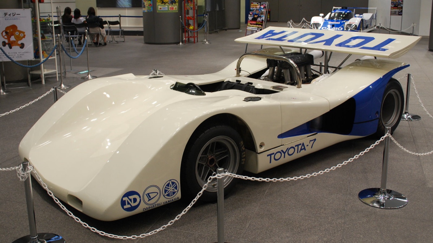 White and blue Toyota 7 open racing car with a tall rear wing and exposed engine, on display behind rope barriers