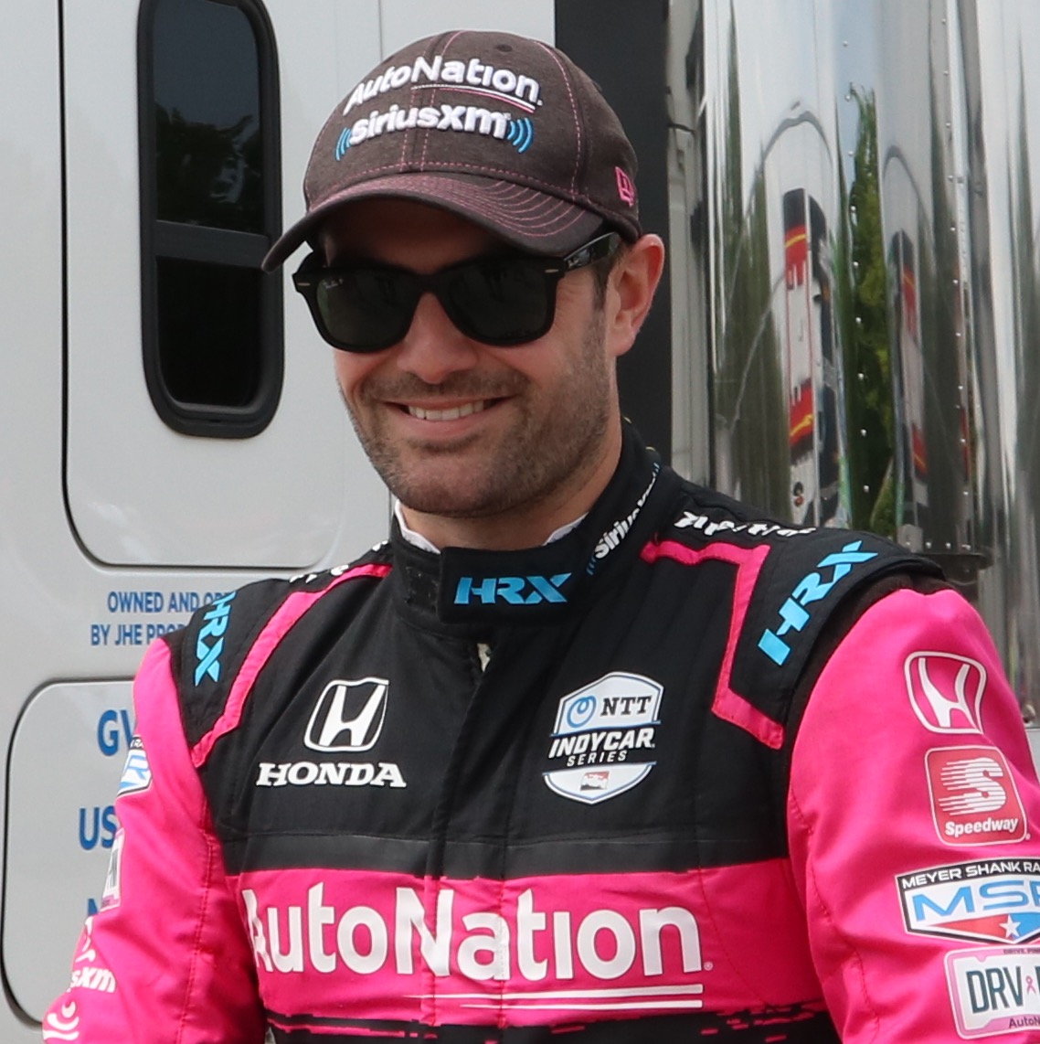 Jack Harvey smiling in a cap and sunglasses, wearing pink and black Meyer Shank Racing overalls
