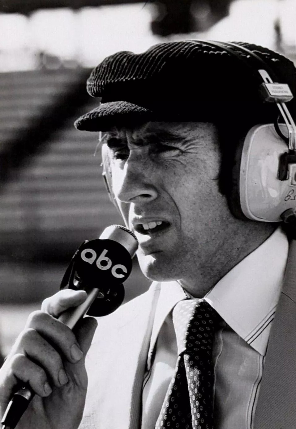 Black and white press photograph of Jackie Stewart in a flat cap and headset, holding an ABC microphone