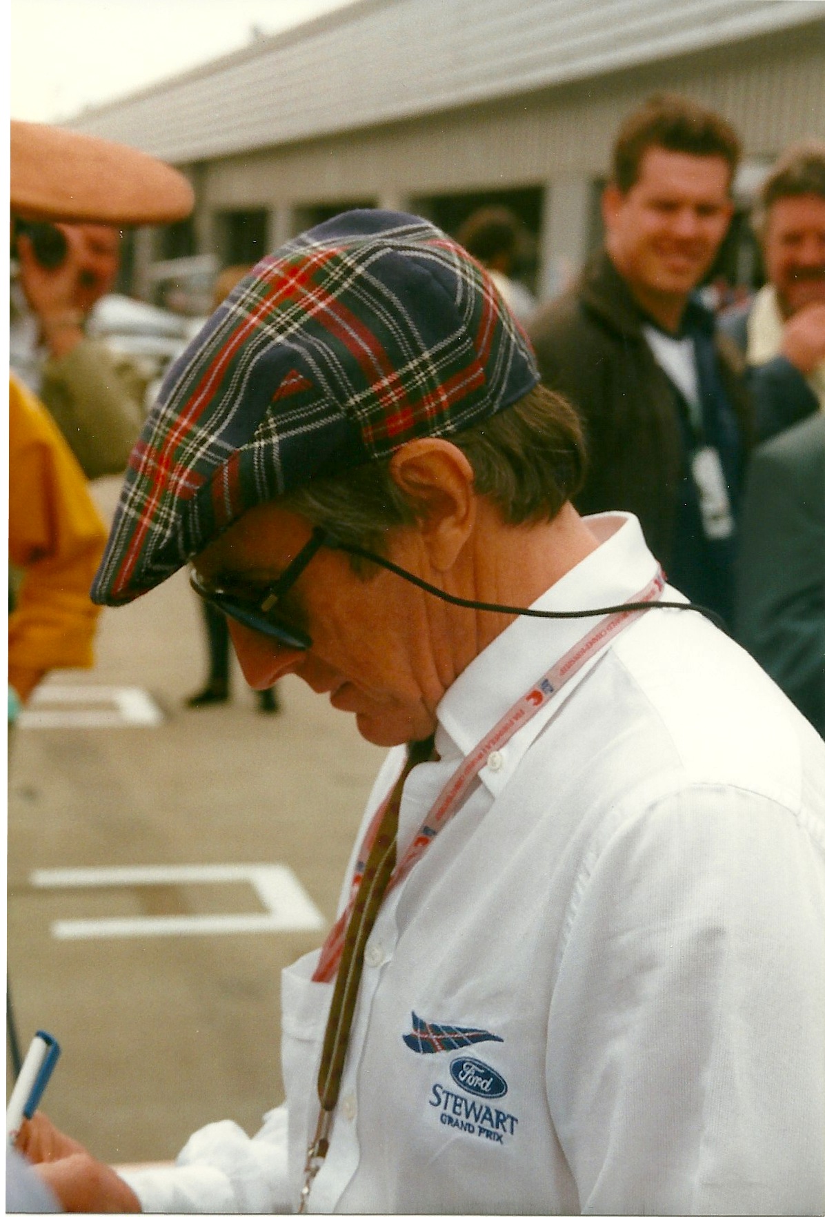 Jackie Stewart in a tartan cap and white Stewart Ford team shirt, looking down at papers in the paddock