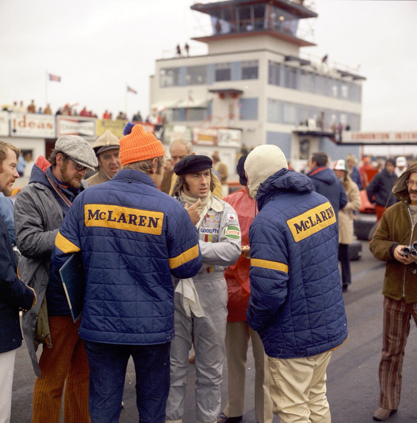 Jackie Stewart in a silver race suit and black cap on the grid at Edmonton, talking to two men in McLaren team jackets
