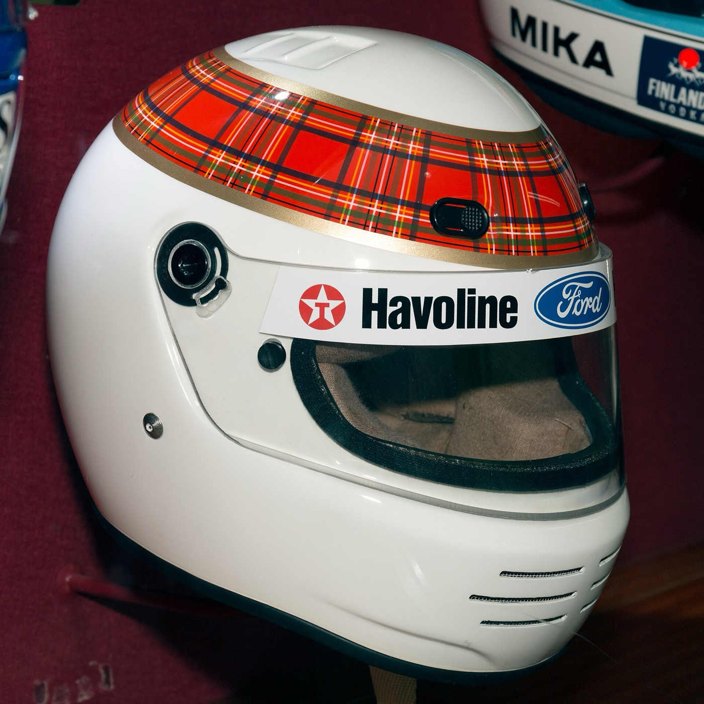 A white full-face helmet of Jackie Stewart’s with a red tartan band round the crown and Havoline and Ford stickers on the visor, in a museum display case