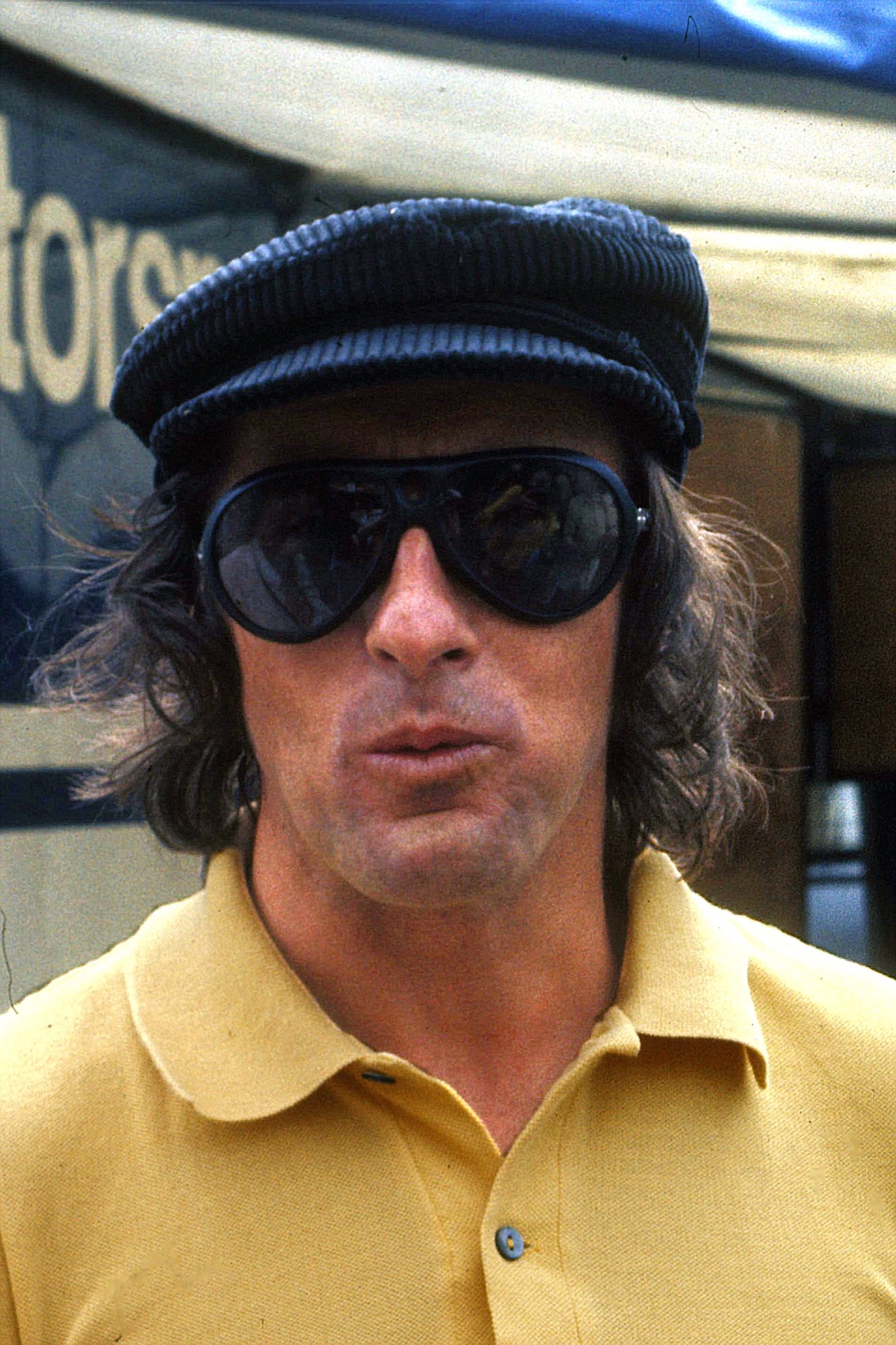 Jackie Stewart in a black corduroy cap, large dark sunglasses and a yellow shirt in the Nürburgring paddock