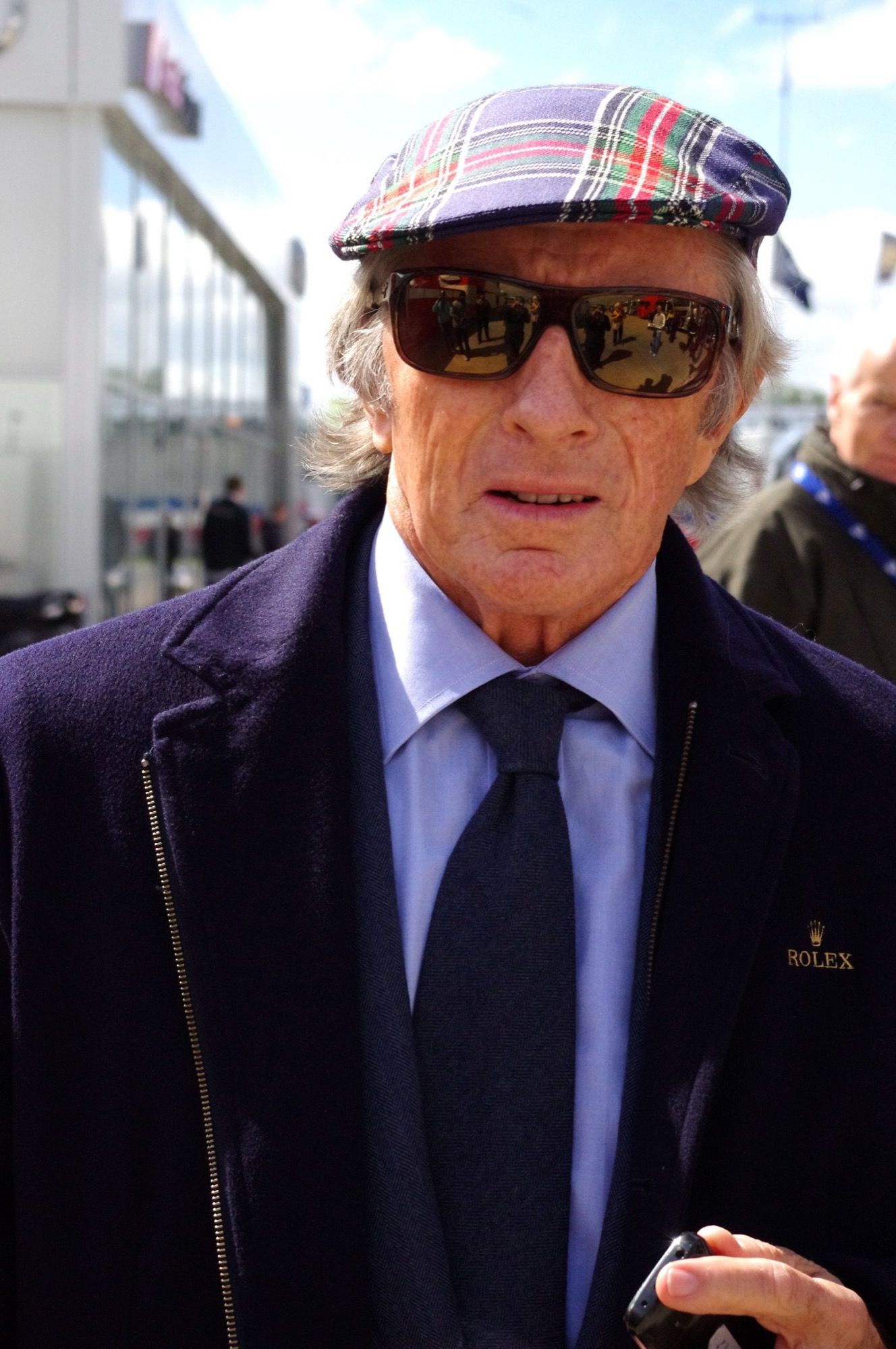 Sir Jackie Stewart in a tartan cap, sunglasses and dark overcoat in the paddock at Silverstone