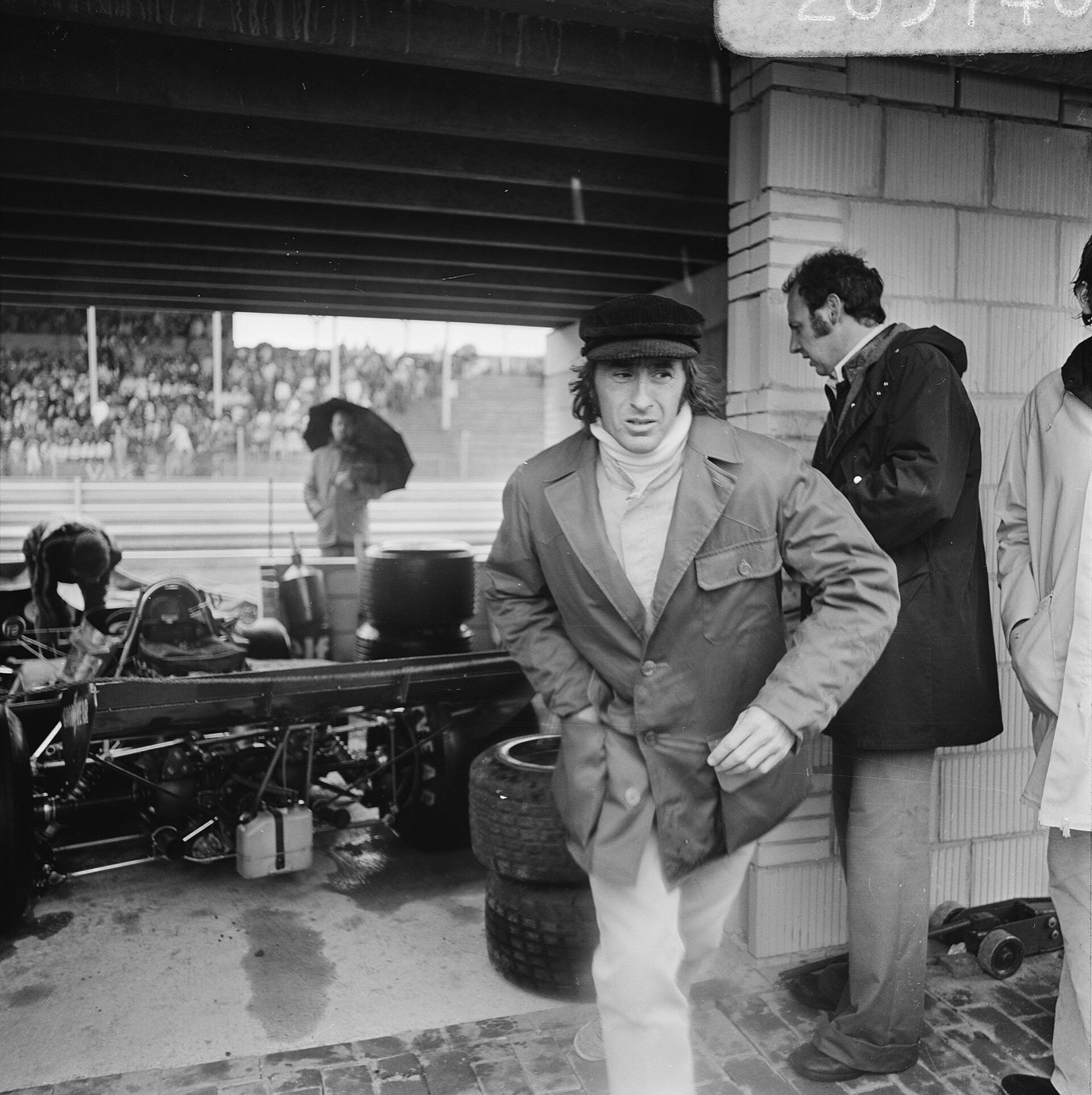 Black and white photograph of Jackie Stewart in a cap and long coat walking past his Tyrrell in the Zandvoort pit lane