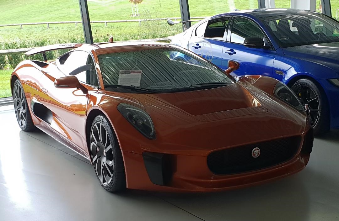 Orange C-X75 hybrid prototype with its rear wing raised, parked beside a blue Jaguar saloon in a museum hall
