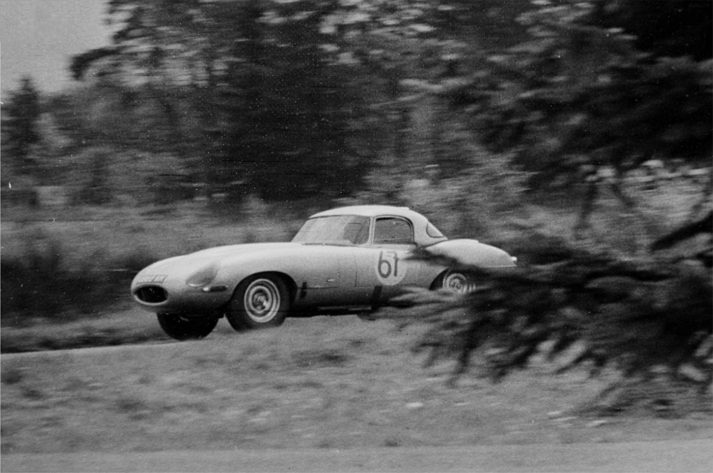 Black and white photograph of a Lightweight E-Type with an aluminium hardtop, race number 61, at speed past trees