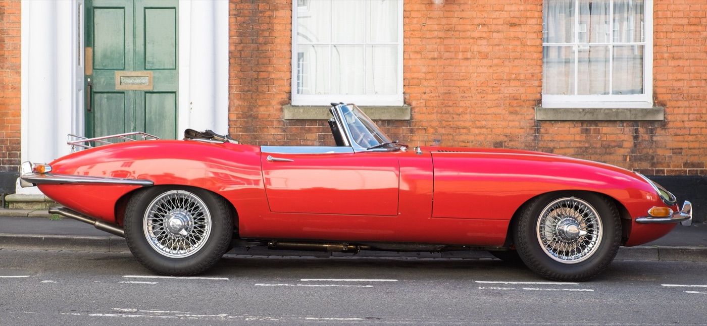 Red Series 1 4.2 E-Type roadster with wire wheels and a boot rack, parked side-on outside a brick terrace