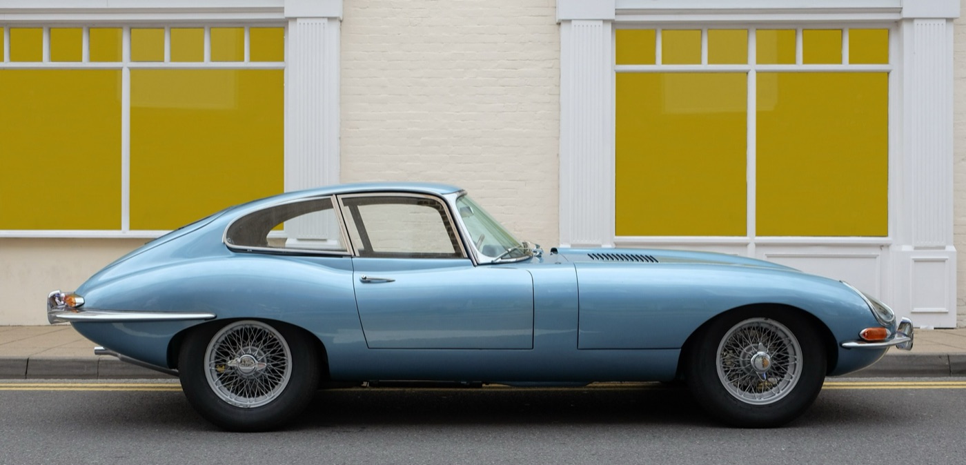 Pale blue Series 1 E-Type fixed-head coupé seen side-on, with wire wheels and covered headlamps