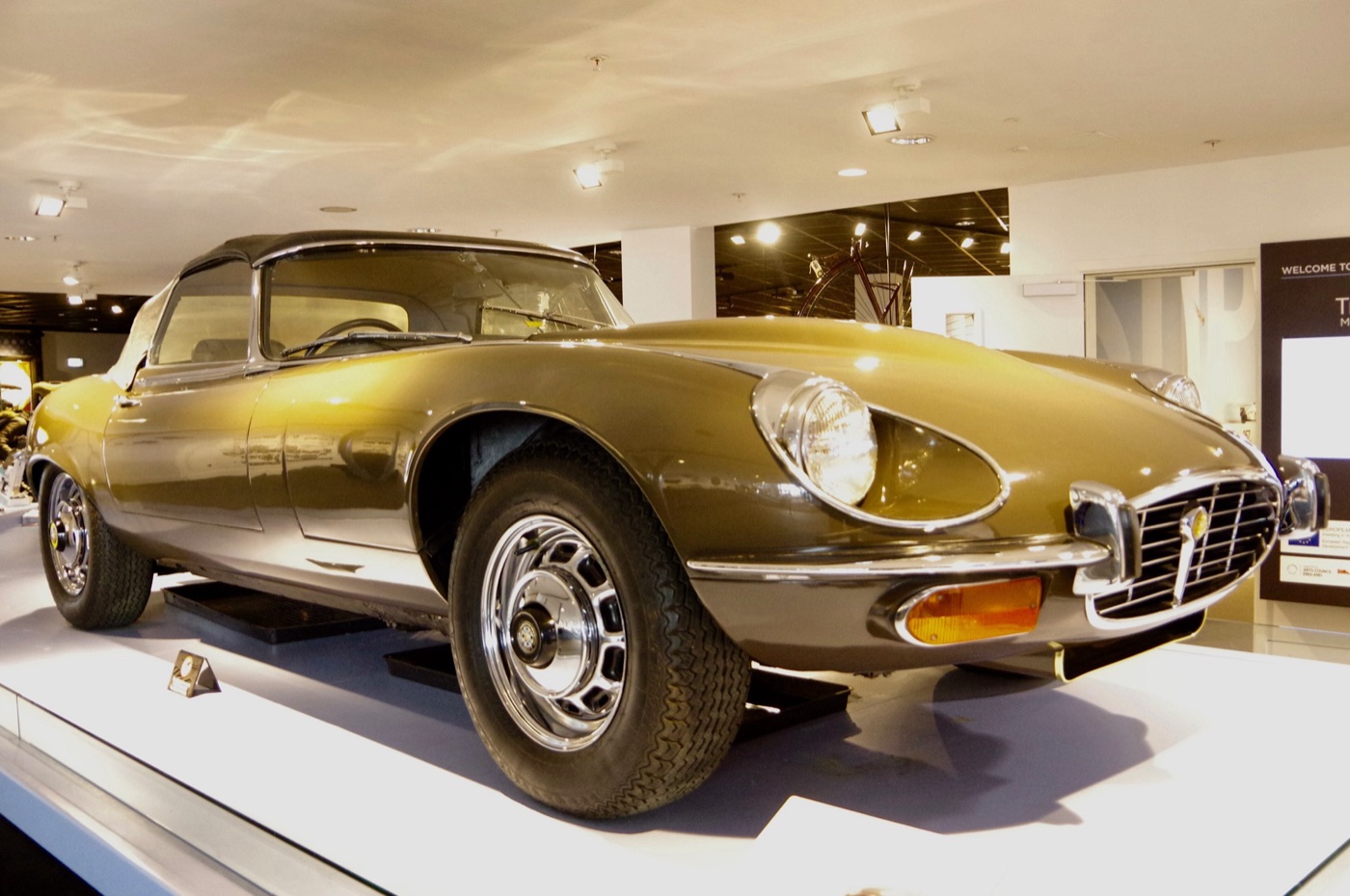 Olive-green Series 3 V12 E-Type roadster with a slatted chrome grille and pressed-steel wheels in a museum hall