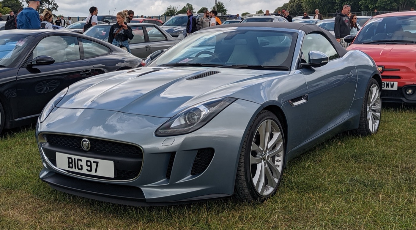 Pale blue-grey 2013 Jaguar F-Type V6 convertible with its black fabric roof up, parked on grass at a car show