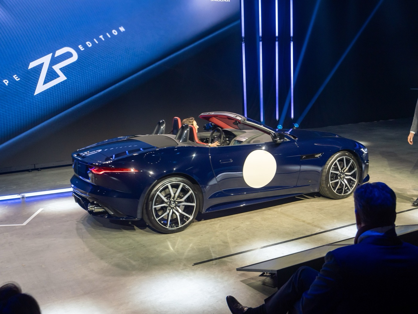 Dark blue Jaguar F-Type ZP Edition convertible with a white racing roundel on the door, roof down, on a lit stage