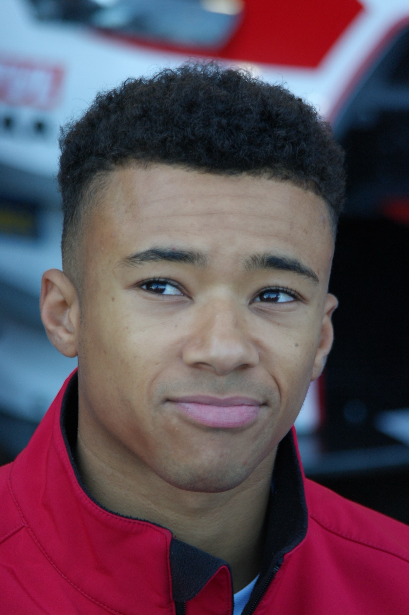 Head and shoulders portrait of Jann Mardenborough in a red team jacket, a race car blurred behind him