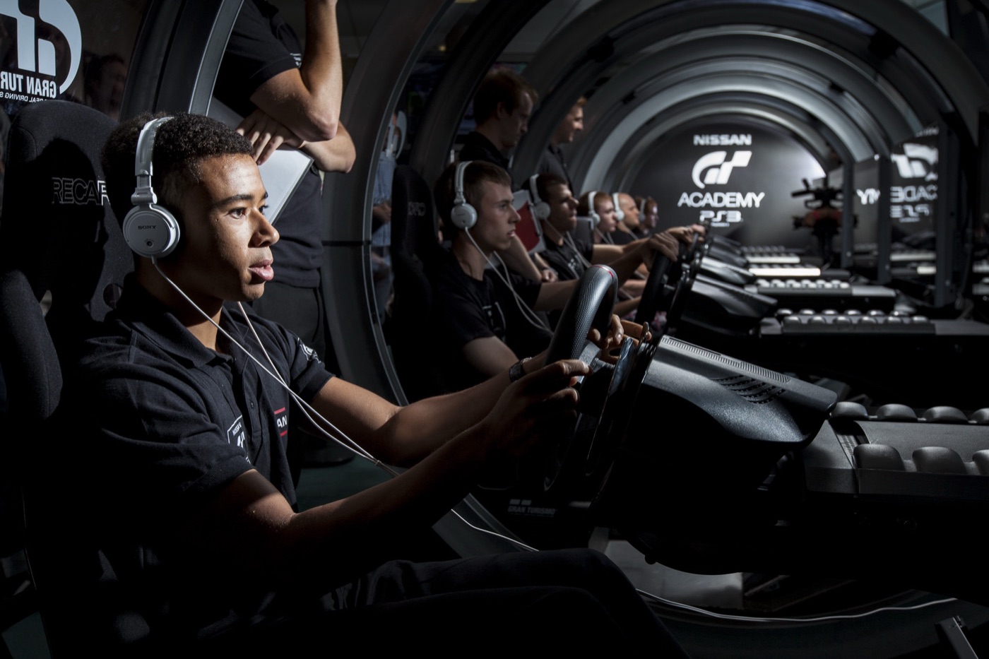 Jann Mardenborough in headphones at a Gran Turismo driving rig, with a row of GT Academy competitors behind him