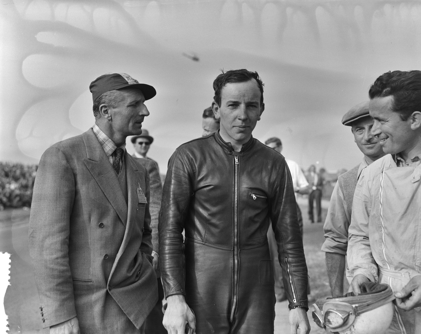 John Surtees in a black leather racing suit after winning the 500cc race at Assen in 1956, with officials and riders around him