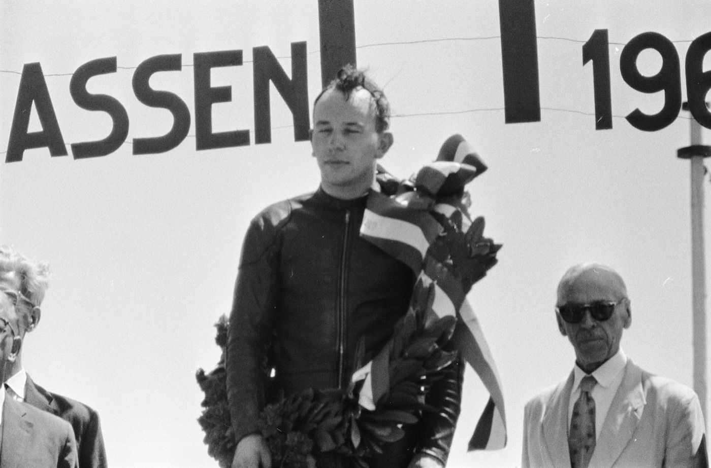 John Surtees on the podium at Assen in 1960 with a laurel wreath and ribbon over his shoulder, under a sign reading Assen