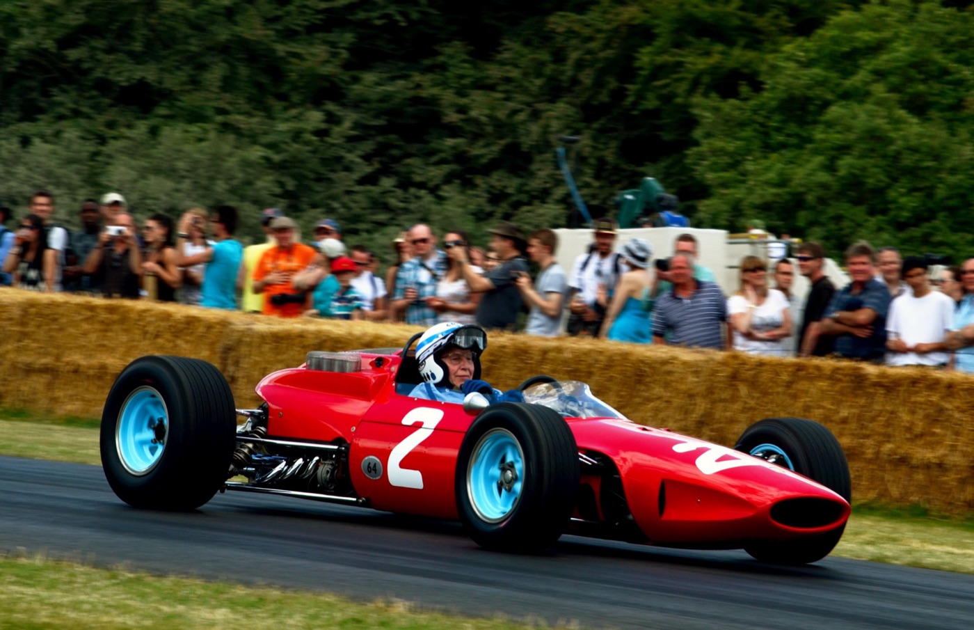John Surtees driving a red Ferrari 158, number 2, past spectators behind straw bales at Goodwood