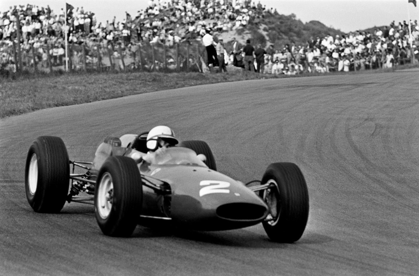 Black and white photograph of John Surtees in the Ferrari 158, number 2, cornering at Zandvoort in front of a crowd on the dunes