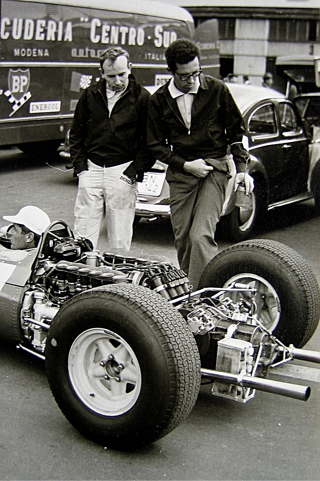 John Surtees and Mauro Forghieri standing behind the flat-12 engine of a Ferrari Formula One car in the paddock, a Scuderia Centro Sud transporter behind