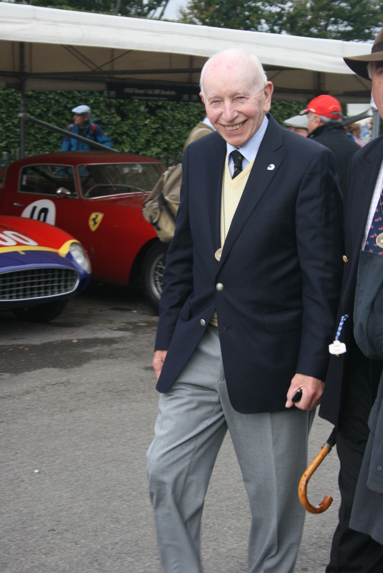 John Surtees in a navy blazer and tie, smiling, walking through the paddock at Goodwood with a Ferrari behind him