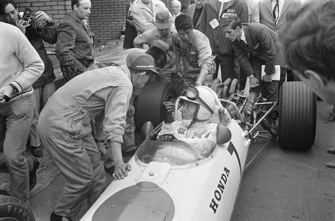 John Surtees sitting in a white Honda Formula One car surrounded by mechanics and photographers
