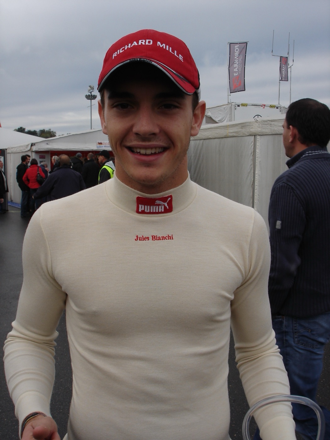 Jules Bianchi in a cream race undershirt with his name stitched on it and a red cap, smiling in a paddock