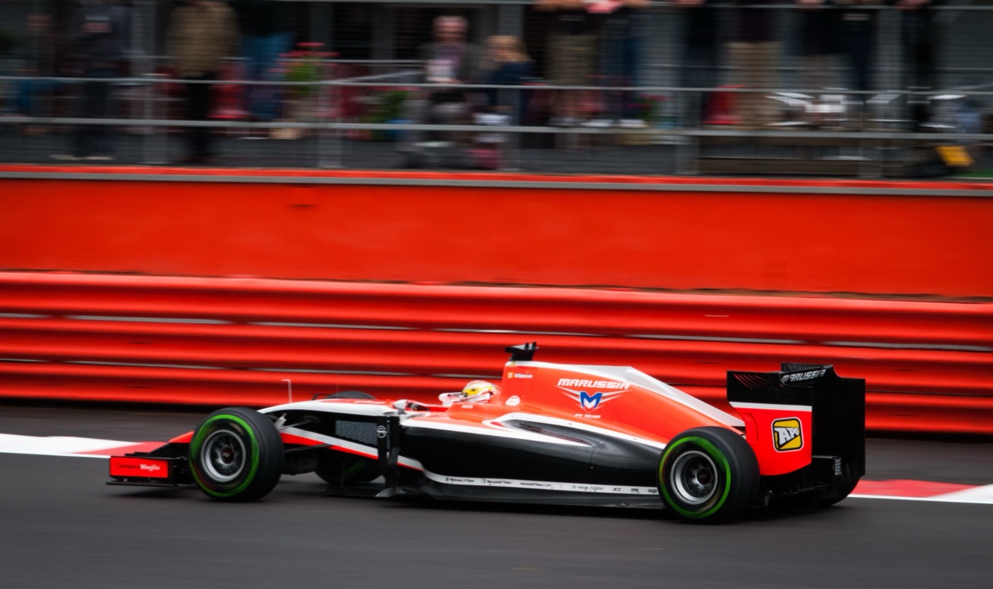 Red and black Marussia MR03 Formula One car at speed past a red barrier, its driver in a white and yellow helmet