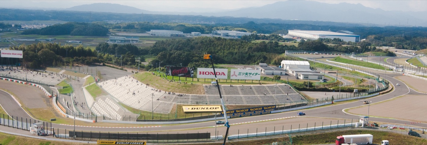 Wide view over the Dunlop Curve at Suzuka Circuit, with grandstands, a Dunlop arch and wooded hills behind