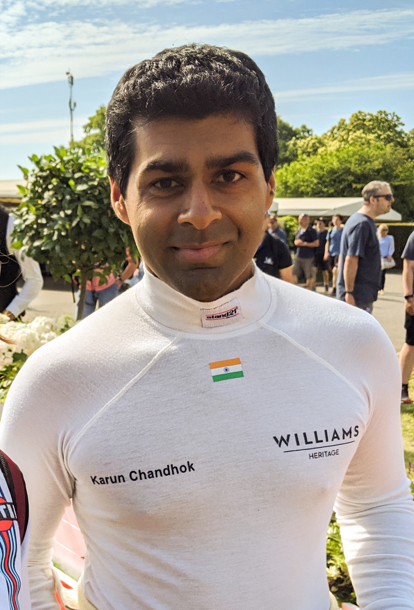 Karun Chandhok smiling, in a white fireproof top with his name, an Indian flag and the Williams Heritage logo, in a crowded paddock