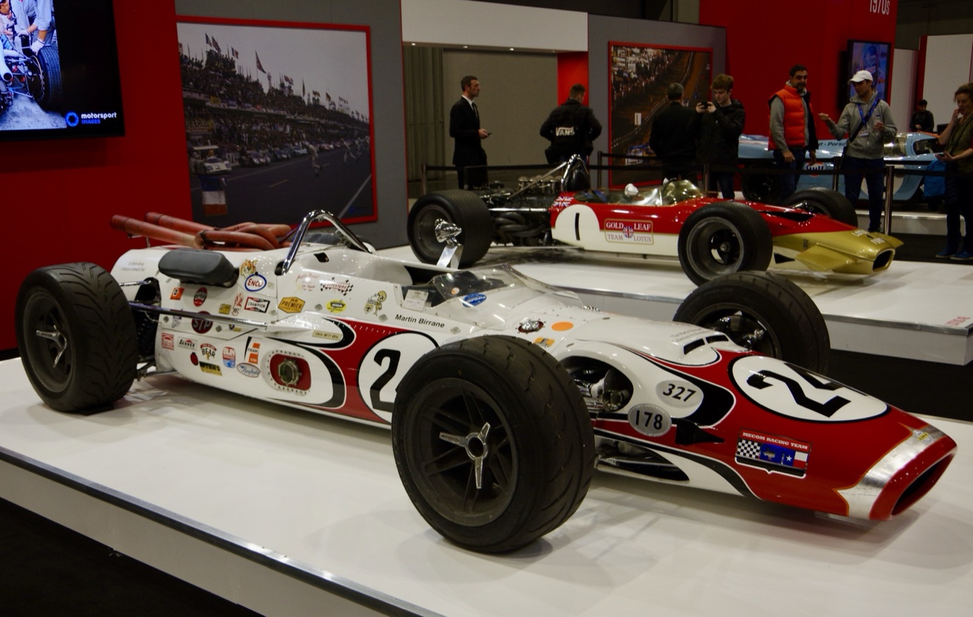 White and red Lola Indianapolis car, number 24, with Martin Birrane’s name on the cockpit side, on a show stand in front of a Gold Leaf Lotus