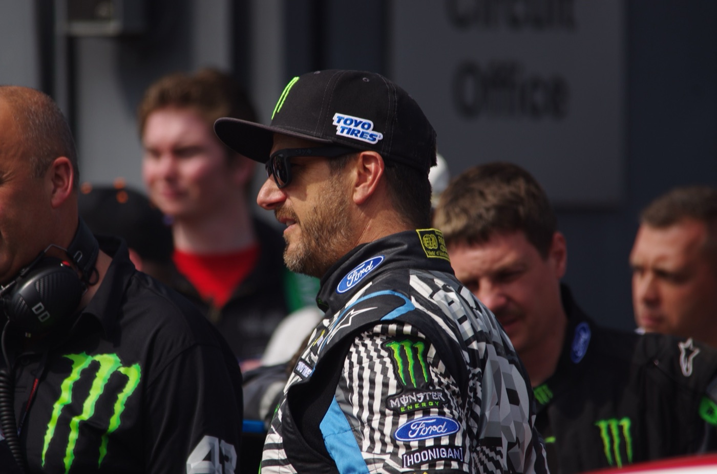 Ken Block in sunglasses, a Toyo Tires cap and a Ford and Monster Energy race suit in a crowded paddock