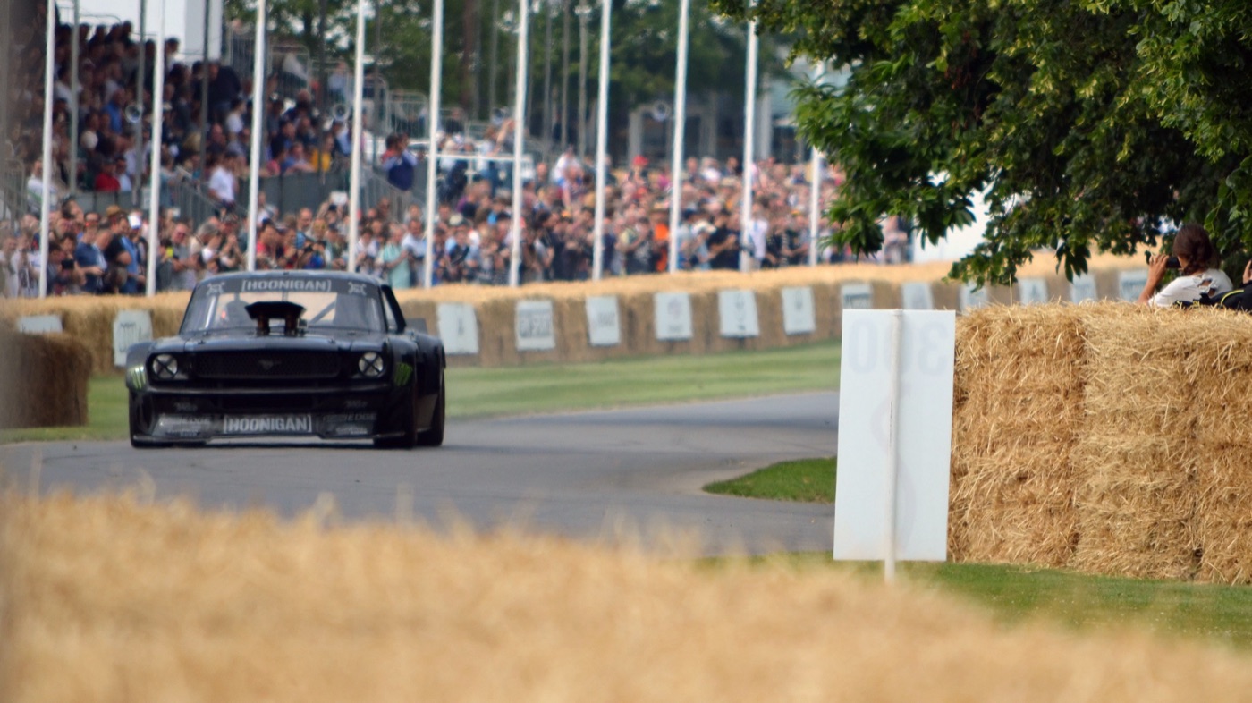 Black 1965 Ford Mustang Hoonicorn with a tall bonnet scoop on the hill-climb course in front of a crowd